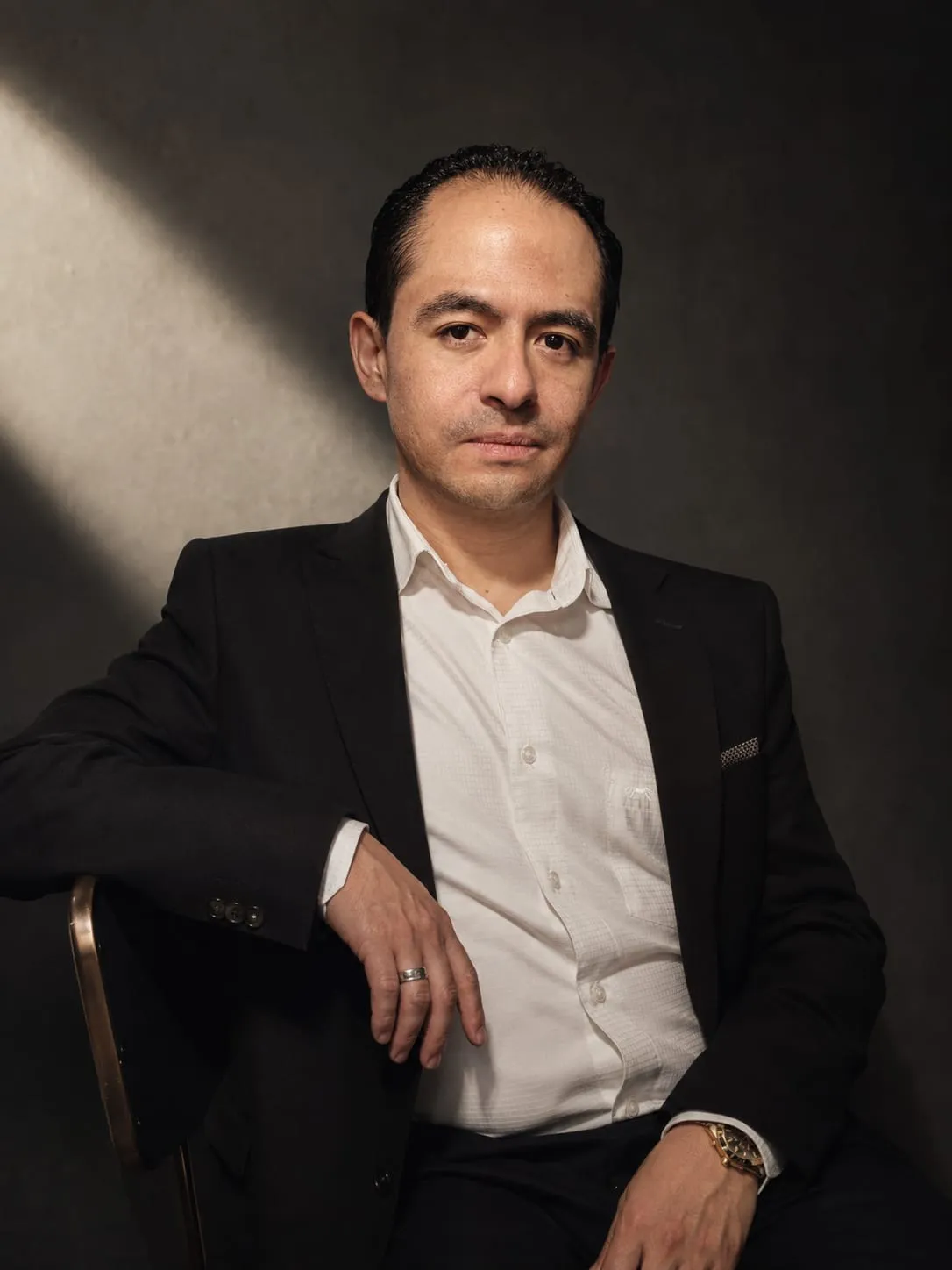 Retrato editorial de Salvador Villarreal sentado, en saco negro y camisa blanca, sobre un fondo oscuro atravesado por una franja de luz.